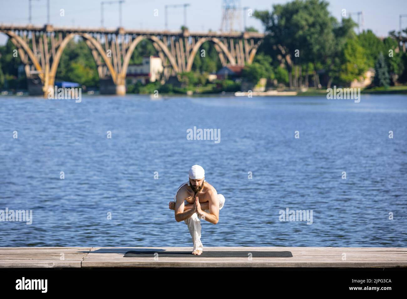 Halten Sie ein Gleichgewicht. Junger Mann praktiziert Yoga am Flussufer bei Sonnenaufgang, im Freien. Konzept der psychischen Gesundheit, Harmonie mit der Natur Stockfoto