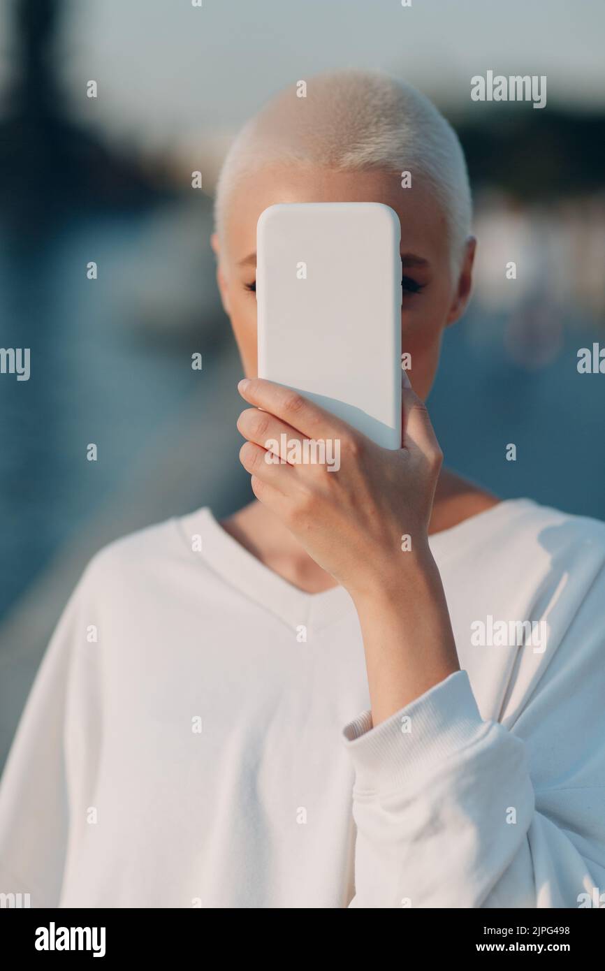 Kurzhaarige Frau, die das Gesicht mit dem Smartphone bedeckt. Konzept für die Datensicherheit, den Datenschutz und die Vertraulichkeit von Mobiltelefonen. Stockfoto