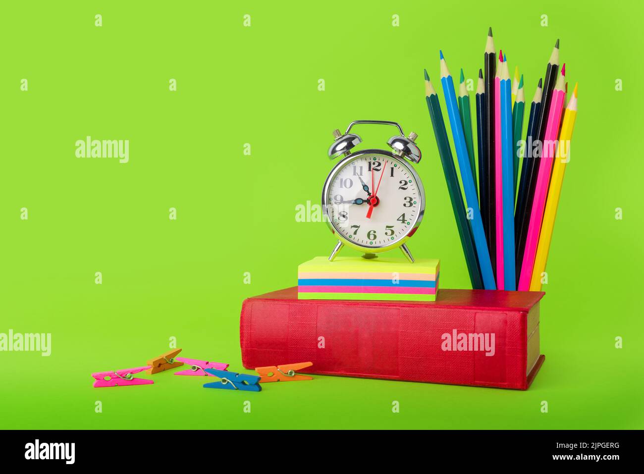 Zurück zur Schule. Eine Uhr neben einem Buch und Buntstiften auf grünem Hintergrund, die das Ende der Ferien symbolisieren. Zeit zum Lernen Stockfoto