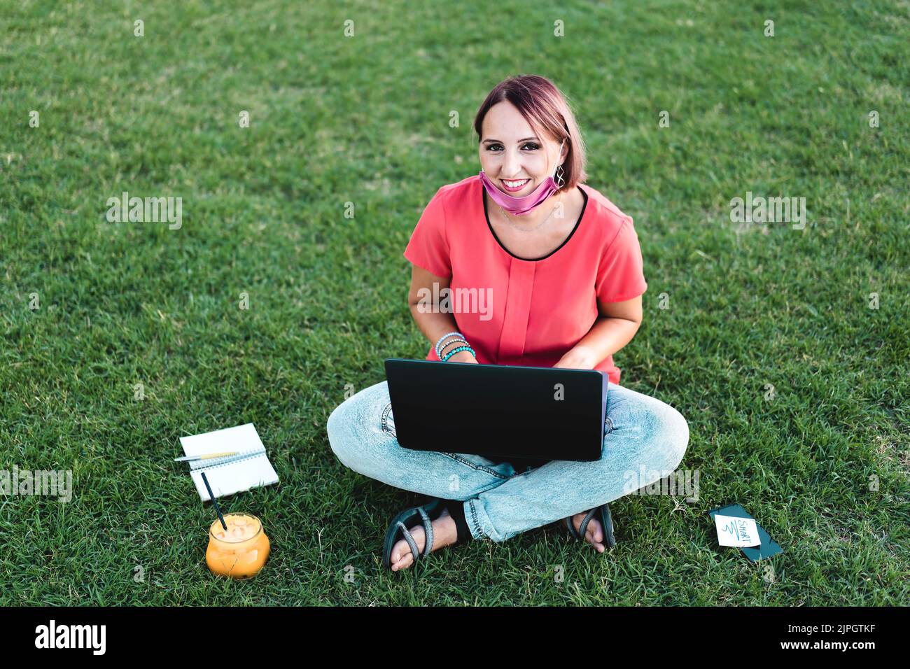 Frau 40 Jahre alt arbeitet im Freien mit Laptop während Coronavirus Outbreak - lächelnde Unternehmerin sitzt auf Rasen mit pc Während Sie Saft trinken Stockfoto