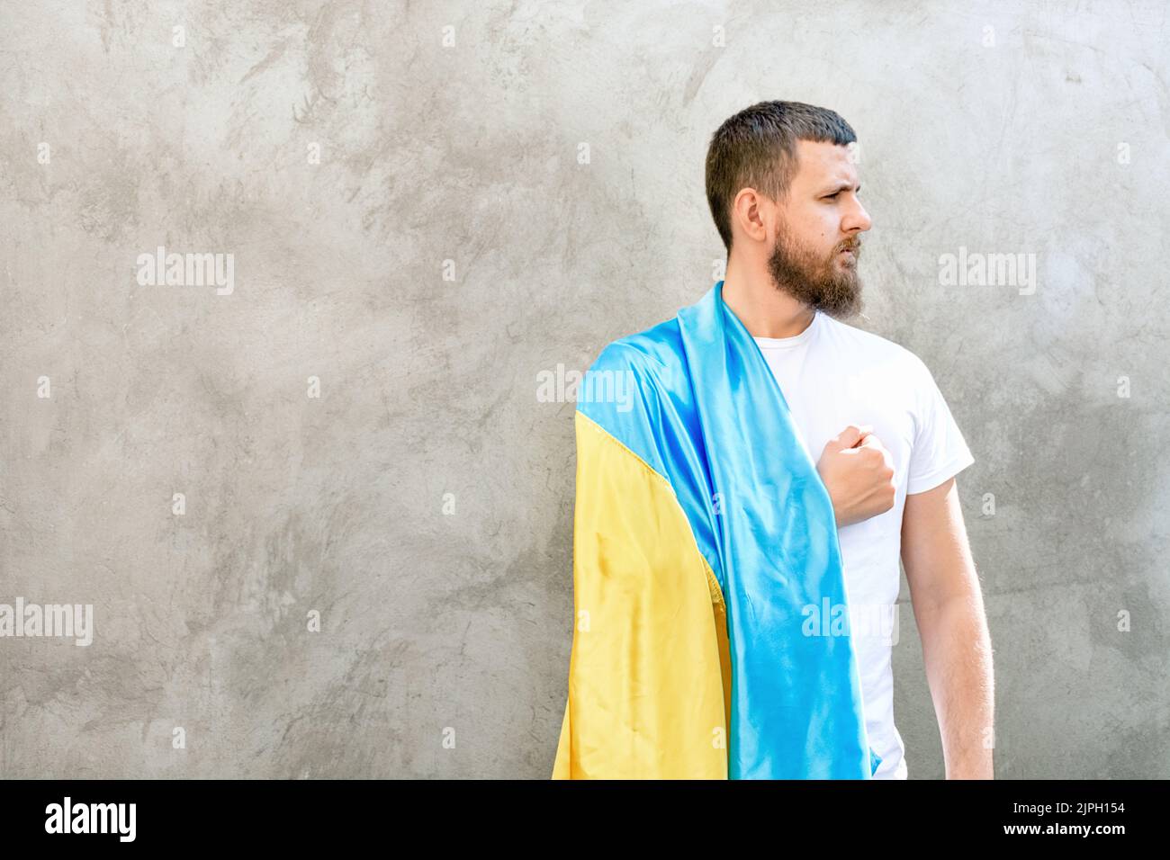 Der Mann hält die Hand über sein Herz und die nationale blau-gelbe Flagge der Ukraine auf seiner Schulter. Typ in weißem T-Shirt und Jeans. Sonniger Tag. Verfassung und Unabhängigkeitstag der Ukraine. Weicher, selektiver Fokus Stockfoto