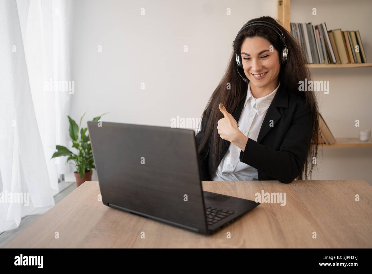 asiatische Geschäftsfrau im Büro, die mit einem Laptop und Kopfhörern arbeitet und den Daumen nach oben zeigt. Selbstbewusste Geschäftsfrau, die Kopfhörer trägt und darüber spricht Stockfoto