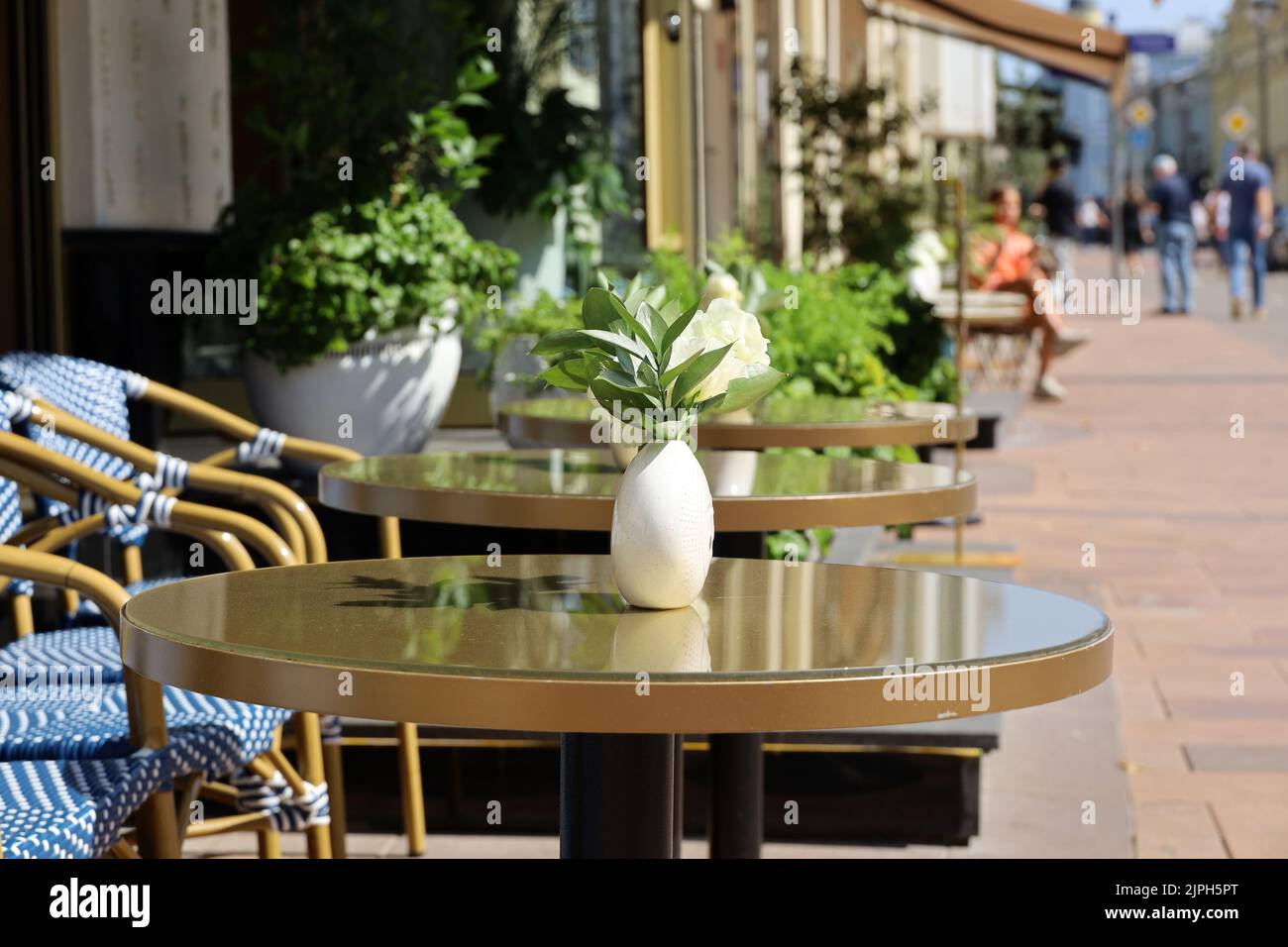 Straßencafé in der Stadt mit leeren Tischen im Freien im Hintergrund von Wanderern. Vasen aus Rosenblumen auf runden Tischen und gemütlichen Stühlen an sonnigen Tagen Stockfoto
