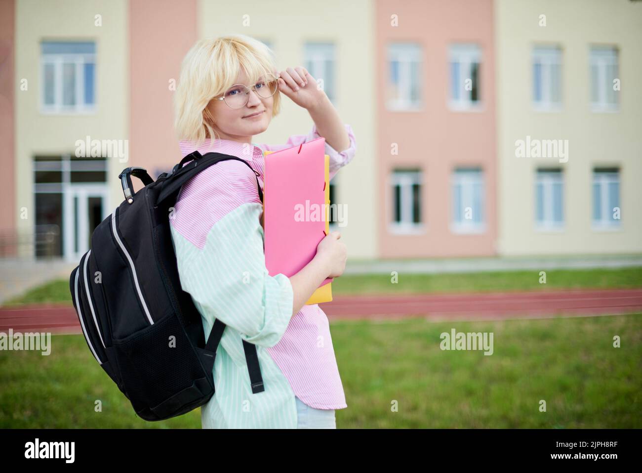 Schülerin im rosa und grünen Hemd mit schwarzem Rucksack und Heften für Notebooks in den Händen zur Schule gehen Stockfoto