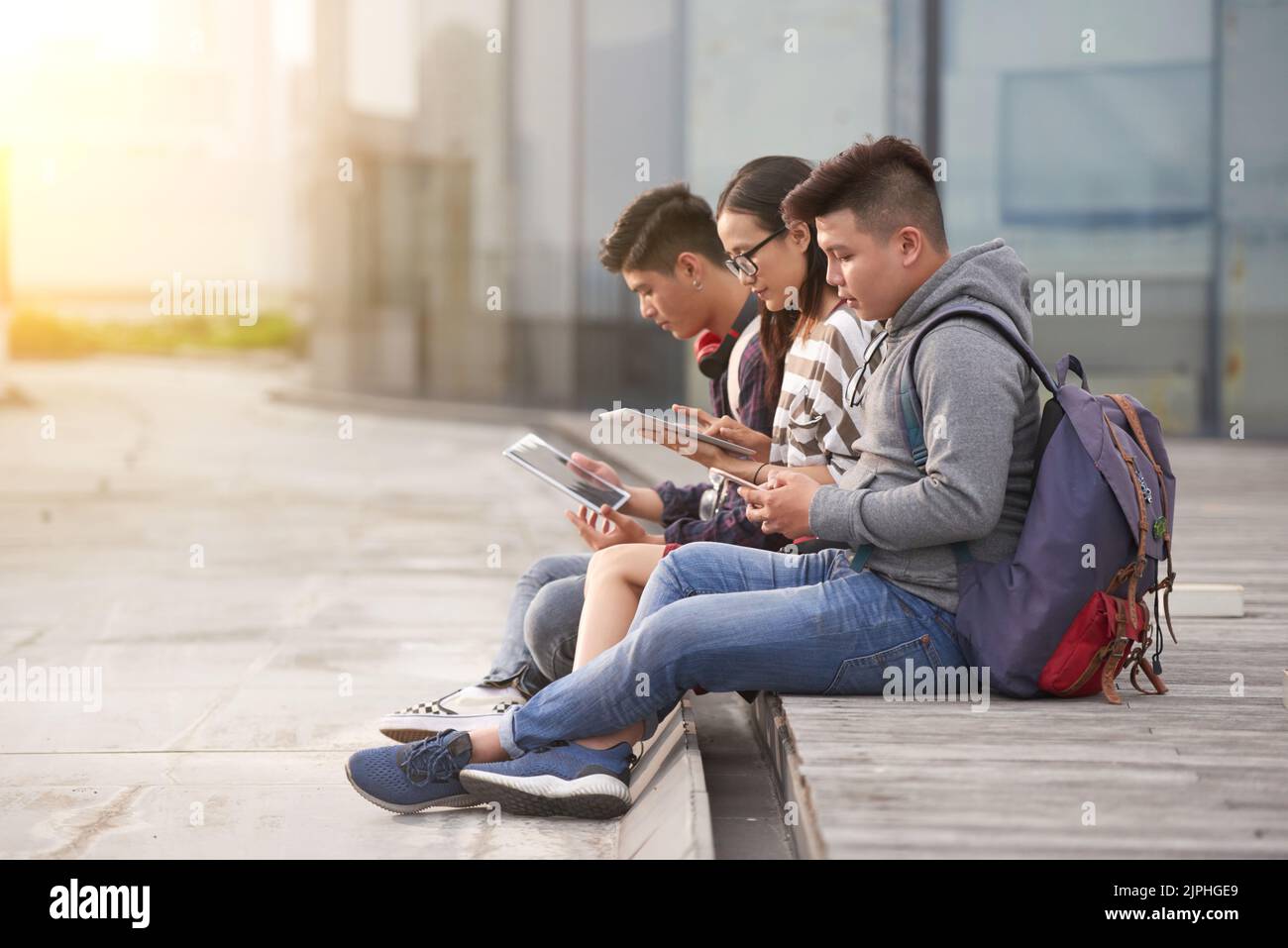Gruppe von asiatischen Studenten in Casualwear sitzen im Freien und surfen Internet auf modernen digitalen Tablets, während sie auf den Beginn des Unterrichts warten, Fassade der modernen Hochschule auf dem Hintergrund Stockfoto