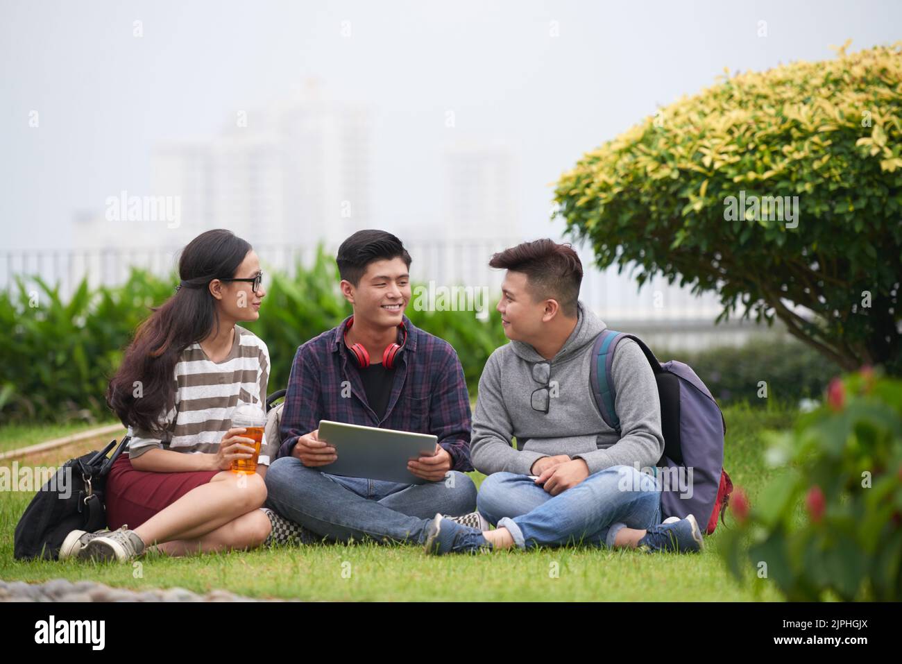 Eine Gruppe von fröhlichen asiatischen Studenten in der Freizeitkleidung versammelten sich im grünen öffentlichen Park und bereiteten sich mit Hilfe eines modernen digitalen Tablets auf die Prüfung vor Stockfoto
