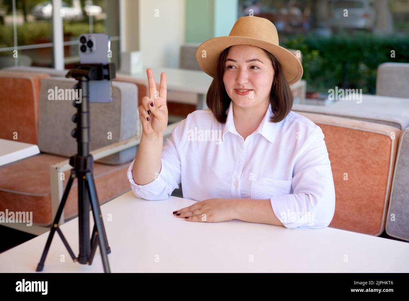 Frau biegt ihre Finger und zählt laut Nummer zwei auf Kamera-Handy. Frau online mit Handy auf Stativ Streaming im Sommer Terrasse Café Stockfoto