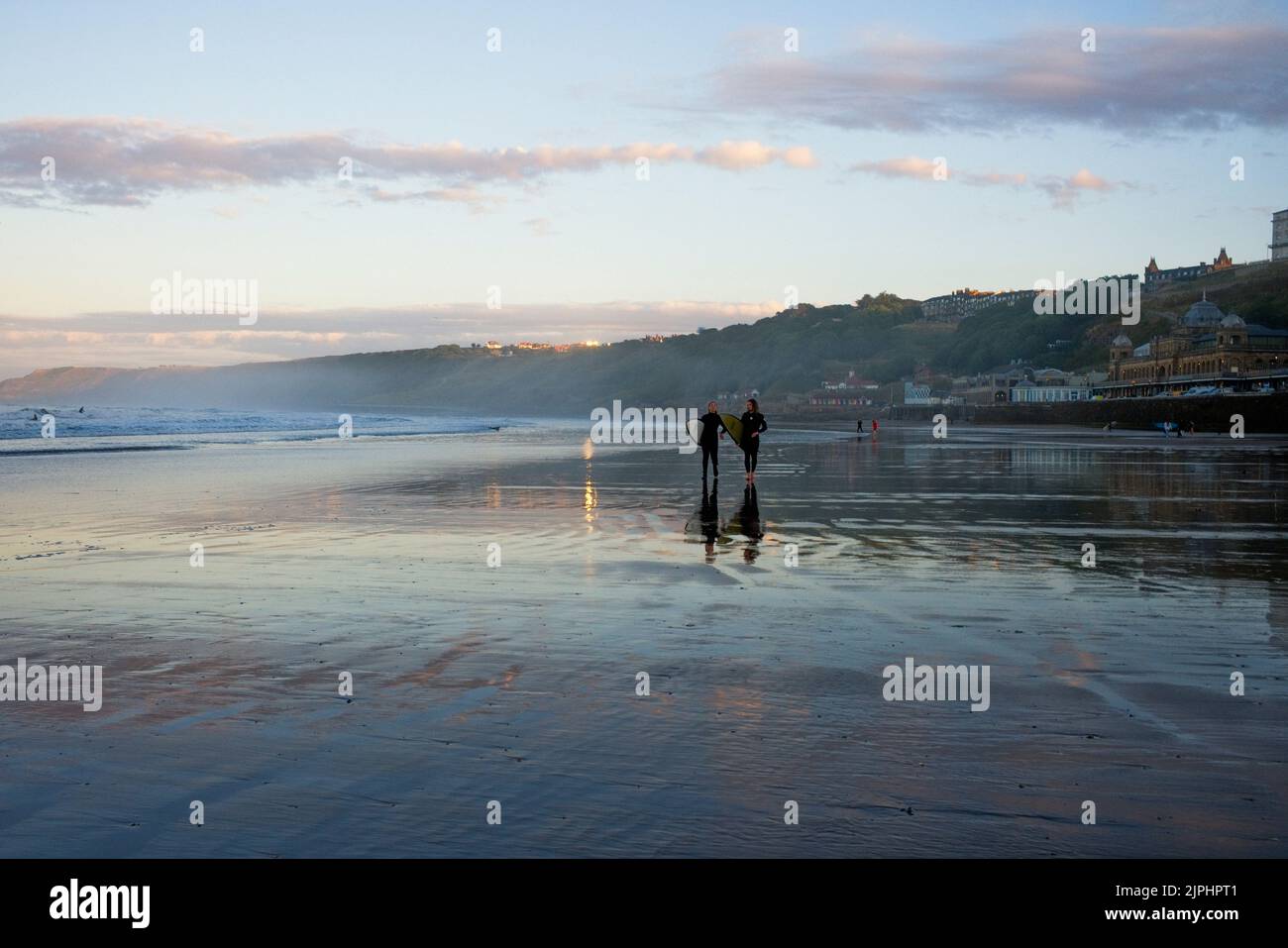 Surfpaar am Scarborough Beach in der Abenddämmerung, das mit anderen Surfern in der Ferne in Richtung Kamera läuft Stockfoto