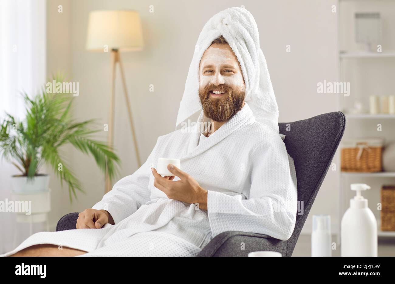 Glücklicher Mann in Bademantel und Handtuch genießen weiße Ton Gesichtsmaske am Tag der Schönheit zu Hause Stockfoto