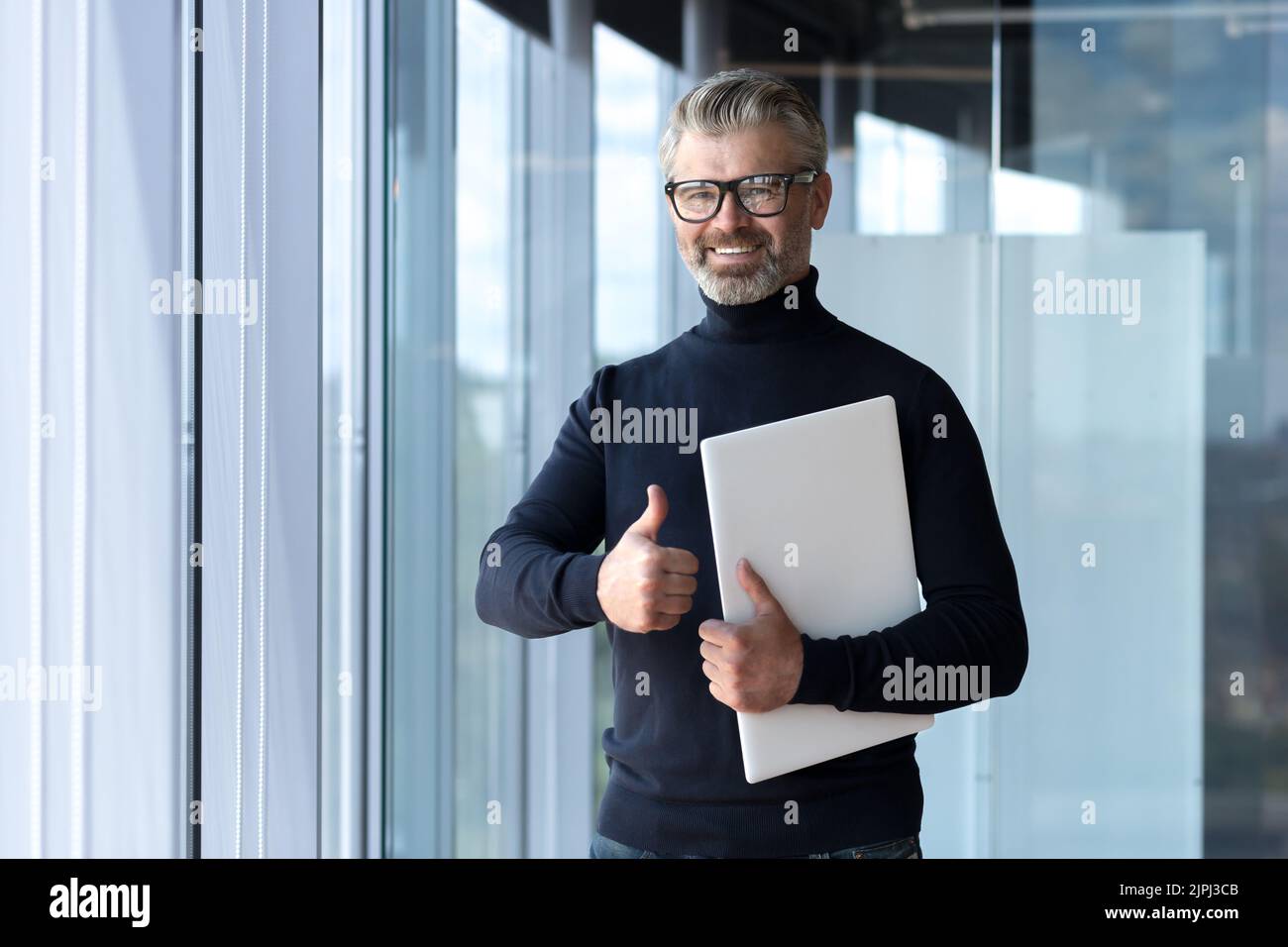 Porträt eines erfolgreichen, erfahrenen und reifen Geschäftsmannes, eines älteren grauhaarigen Mannes, der im Bürogebäude arbeitet, eines Finanziers, der lächelt und mit dem Laptop den Daumen nach oben zeigt Stockfoto