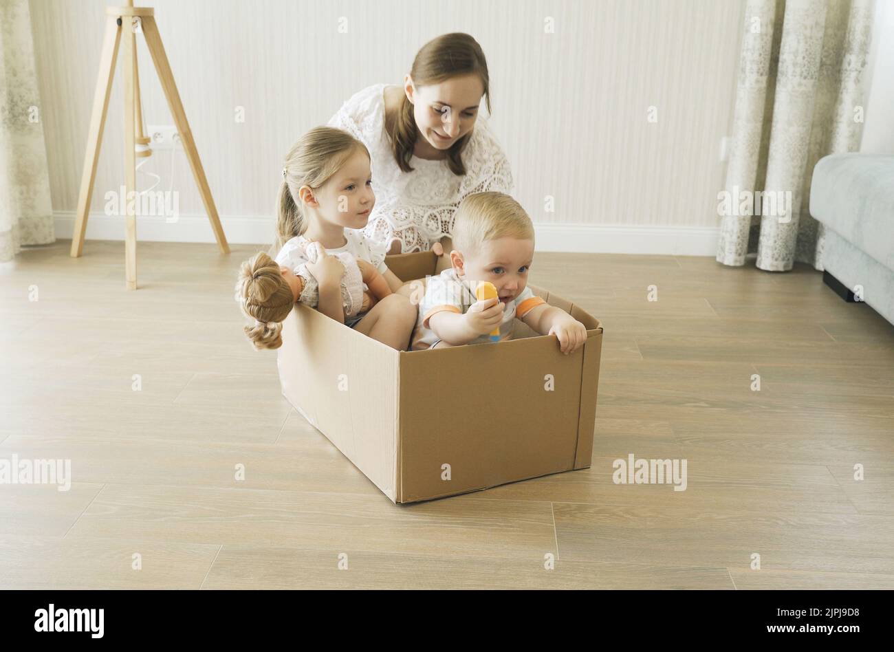 Mama rollt einen fröhlichen Sohn und eine Tochter in einem Pappkarton um den Raum. Spiele mit Kindern. Stockfoto