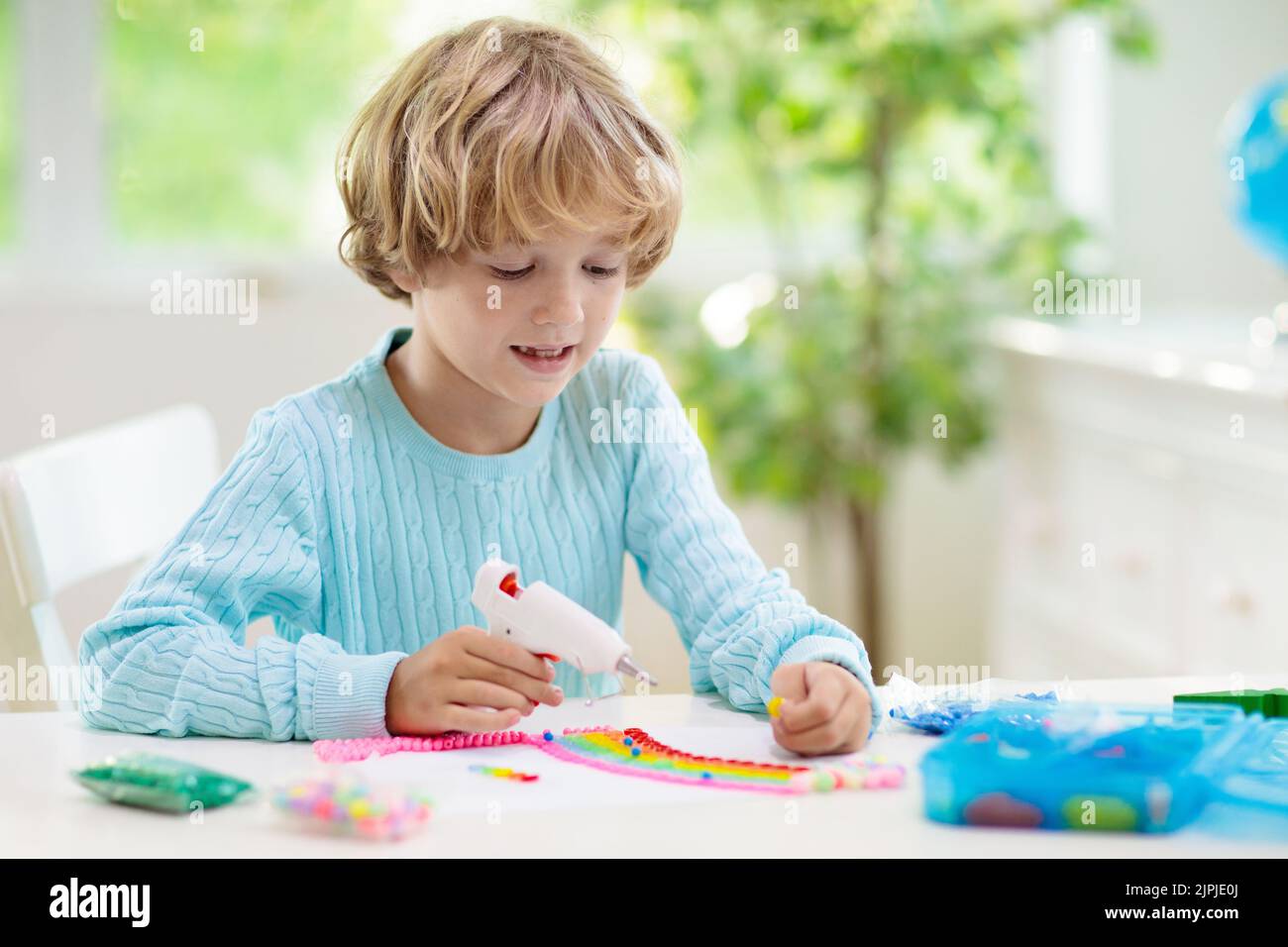 Basteln für Kinder. Kind mit Heißklebepistole. Kreativer kleiner Junge, der Knöpfe und Perlen im Regenbogen-Design klebt. Kunst und Kreativität Klasse in der Schule Stockfoto