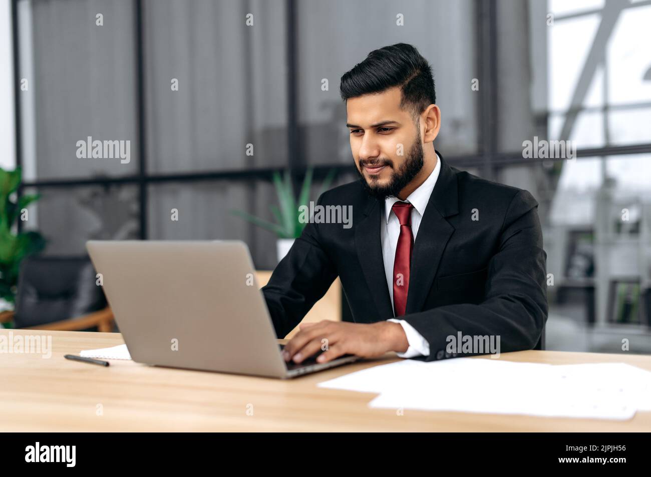 Fokussierter, intelligenter, selbstbewusster arabischer oder indischer erfolgreicher Geschäftsmann, firmenchef, im formellen Anzug, sitzen mit Laptop am Schreibtisch im modernen kreativen Büro, konzentrierter Blick auf den Laptop-Bildschirm Stockfoto
