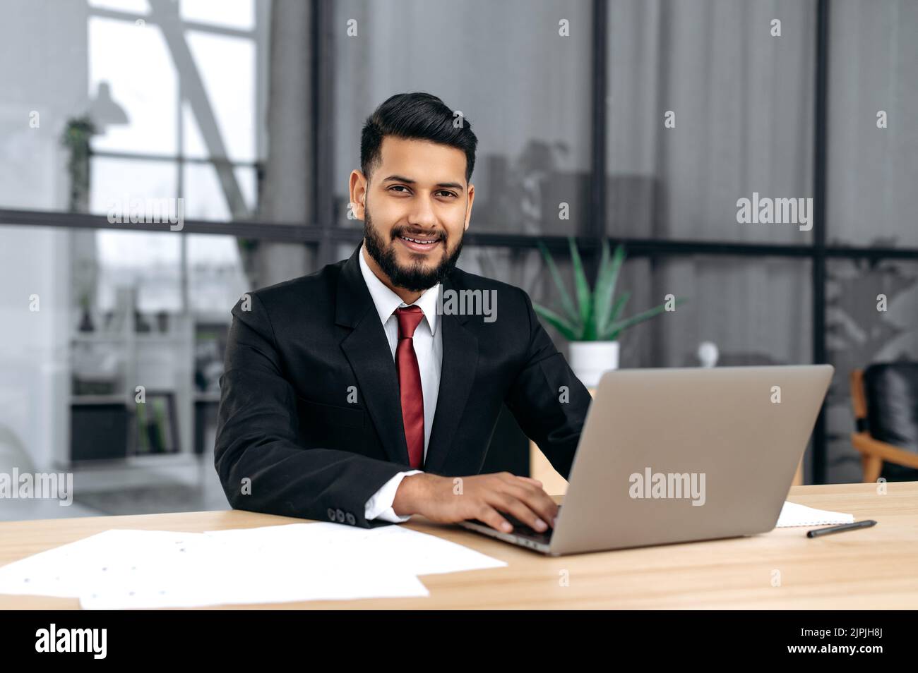 Porträt eines attraktiven eleganten selbstbewussten arabischen oder indischen erfolgreichen Geschäftsmannes, Unternehmers, Rechtsanwalts, in formellen Anzug, sitzen am Schreibtisch mit Laptop in modernen kreativen Büro, schaut auf die Kamera, lächeln Stockfoto