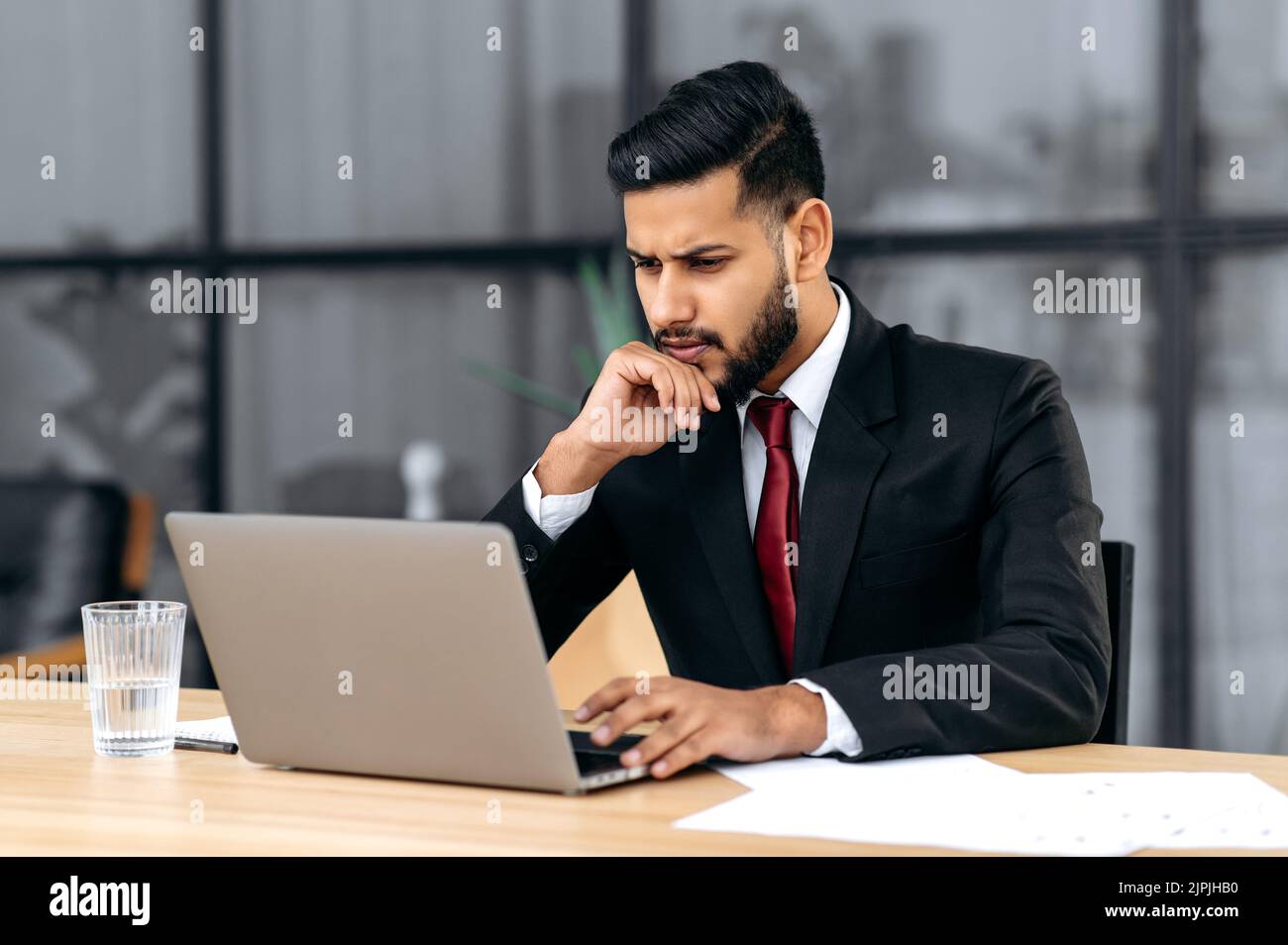 Verwirrt verwirrt verwirrt verwirrt ist der junge indische oder arabische Geschäftsmann, Unternehmer, in einem Anzug, sitzend in seinem modernen Büro, fokussierter Blick auf den Laptop-Bildschirm, unerwartete Nachrichten oder Nachrichten zu sehen, unter Stress Stockfoto