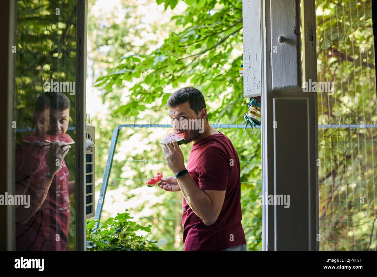 Mann, der saftige rote Wassermelone auf dem Balkon isst. Das Konzept von Sommer und köstlichem Gemüse. Vorderansicht. Stockfoto