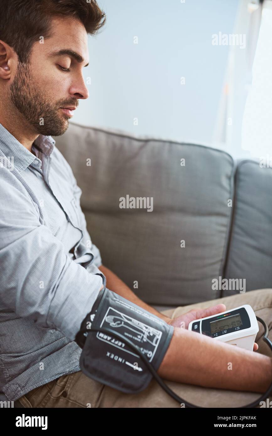 Ein fokussierter junger Mann, der seinen Blutdruck mit einem Blutdruckmessgerät ablesen konnte, während er zu Hause auf einer Couch saß Stockfoto