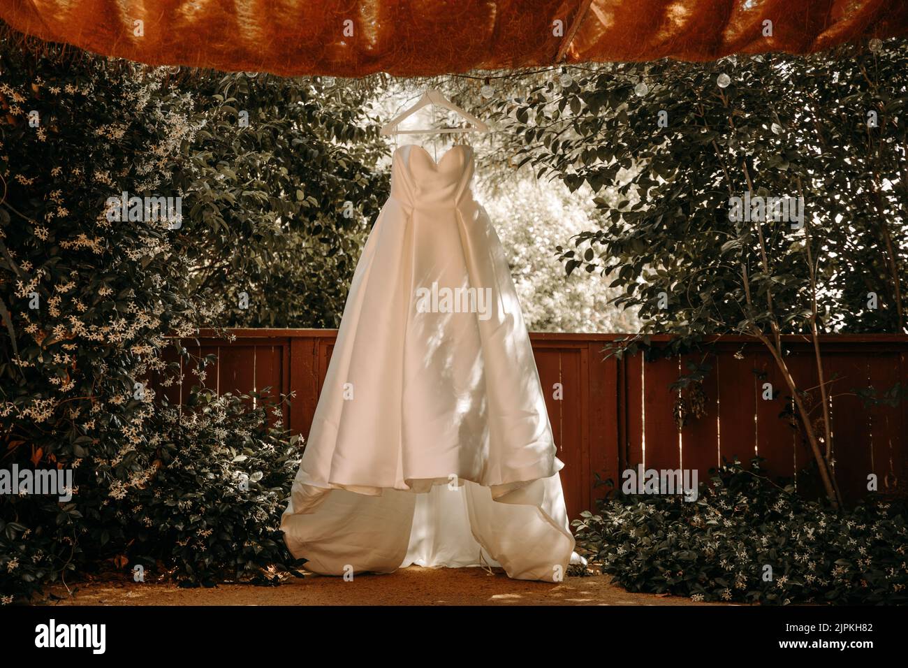 Schönes Hochzeitskleid mit Falten und Schwanz hängen auf weißen Kleiderbügel im Freien. Ballkleid im Garten. Stockfoto