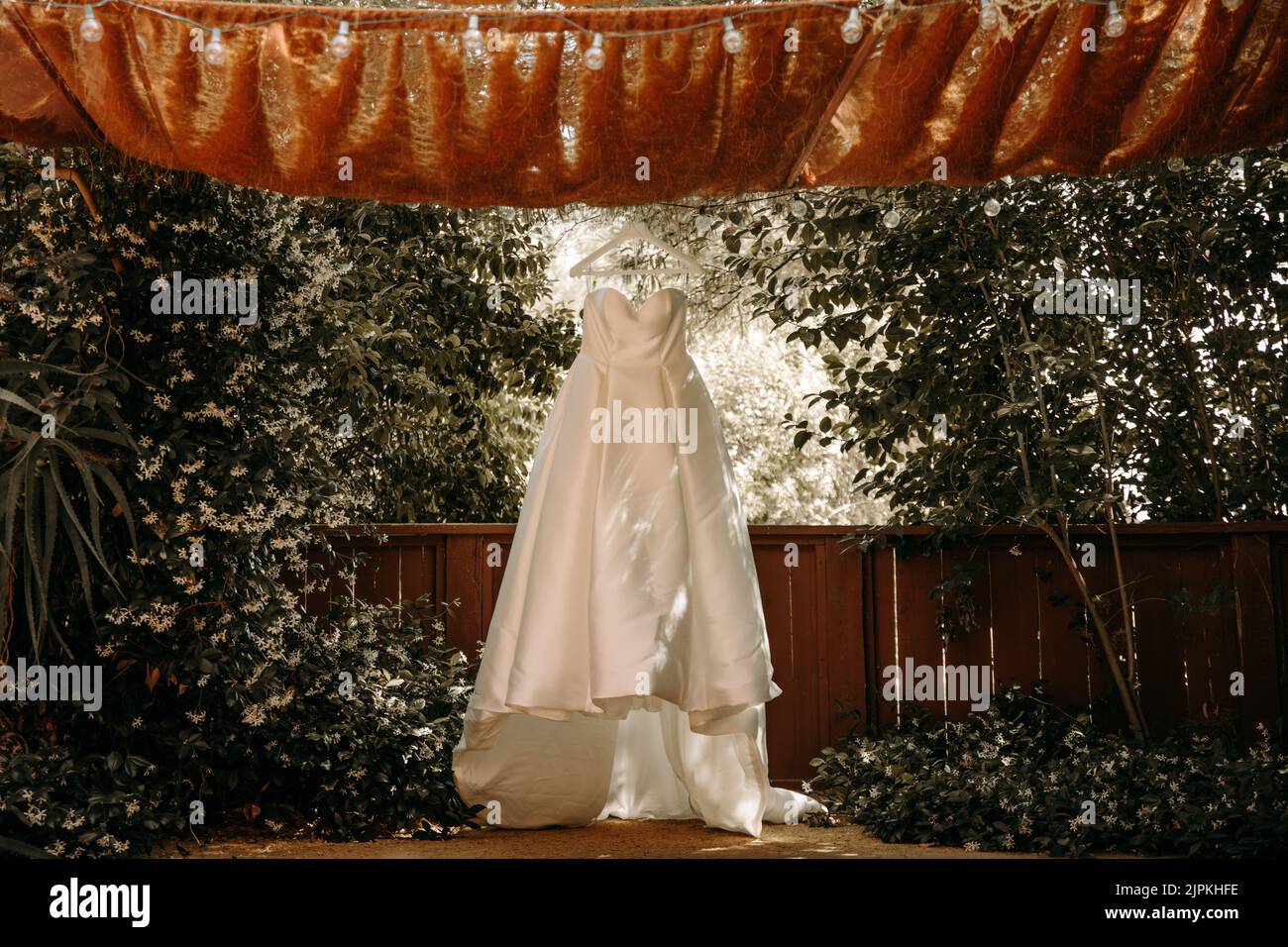 Schönes Hochzeitskleid mit Falten und Schwanz hängen auf weißen Kleiderbügel im Freien. Ballkleid im Garten. Stockfoto