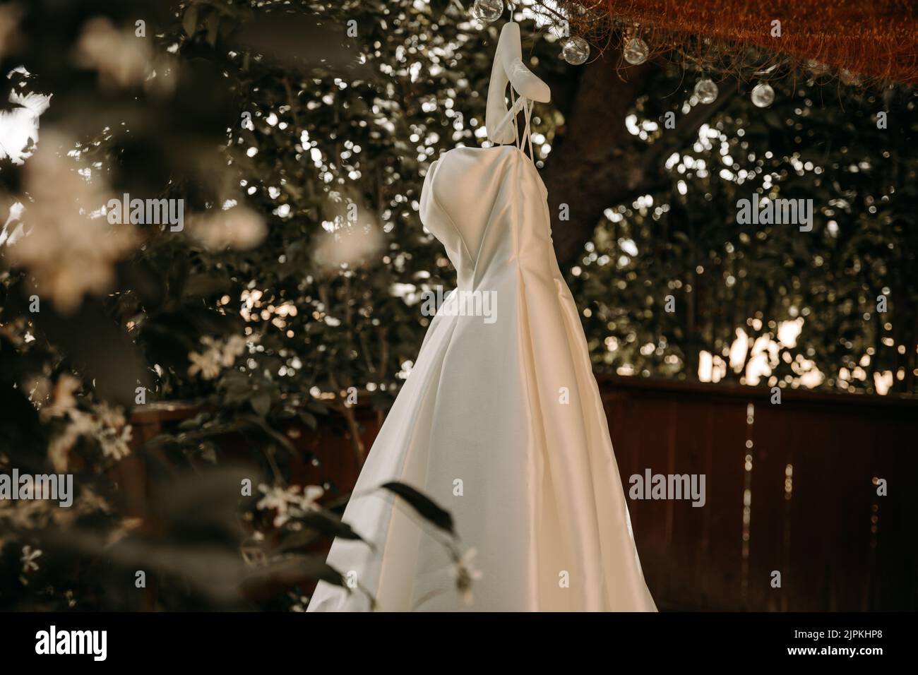 Schönes Hochzeitskleid mit Falten und Schwanz hängen auf weißen Kleiderbügel im Freien. Ballkleid im Garten. Stockfoto