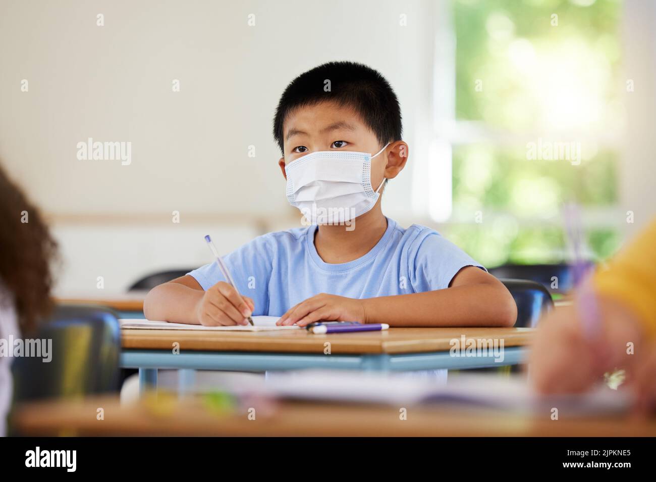 . Gesichtsmaske zum Schutz vor Covid während einer Pandemie, kreative Schüler lernen in der Klasse und schreiben in Notizbuch im Klassenzimmer. Junge, der bei einem sitzt Stockfoto