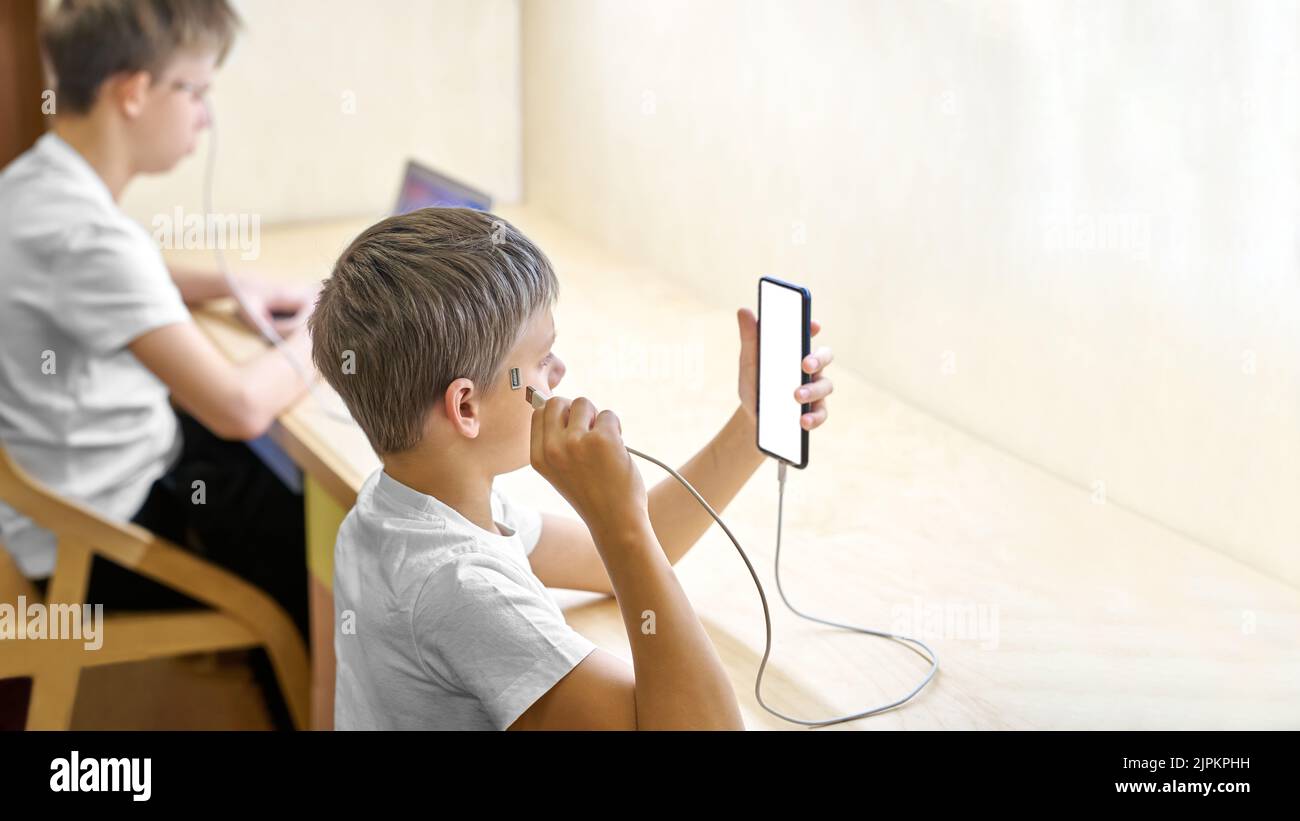 Schüler nutzen elektronische Geräte, die direkt an den Kopf angeschlossen sind. Der jüngere Bruder verbindet das Kabel mit dem Smartphone, während der ältere Bruder über den Laptop spielt Stockfoto