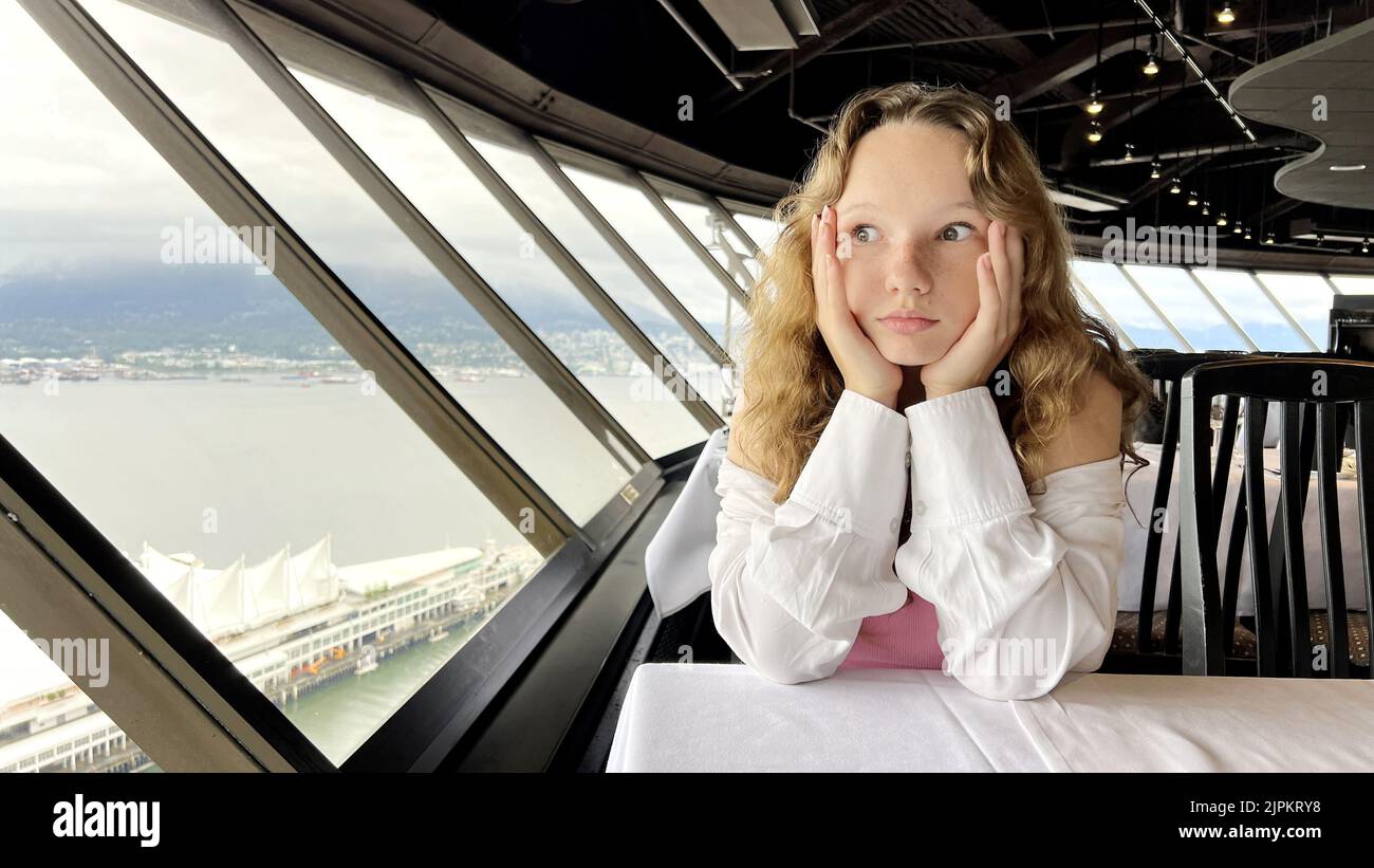 Ein junges Mädchen im Teenageralter sitzt am Fenster eines sich drehenden Restaurants und ist traurig, dass sie ihre Hand neben ihrem Gesicht traurig ist und sie sich die Sehenswürdigkeiten Top of Vancouver Revolving Restaurant ansieht. 4K Stockfoto