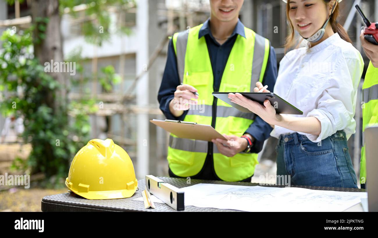 Schöne attraktive asiatische Architektin überprüft einige Details auf Zwischenablage während des Treffens mit einem Team von Bauingenieur auf der Baustelle. Stockfoto