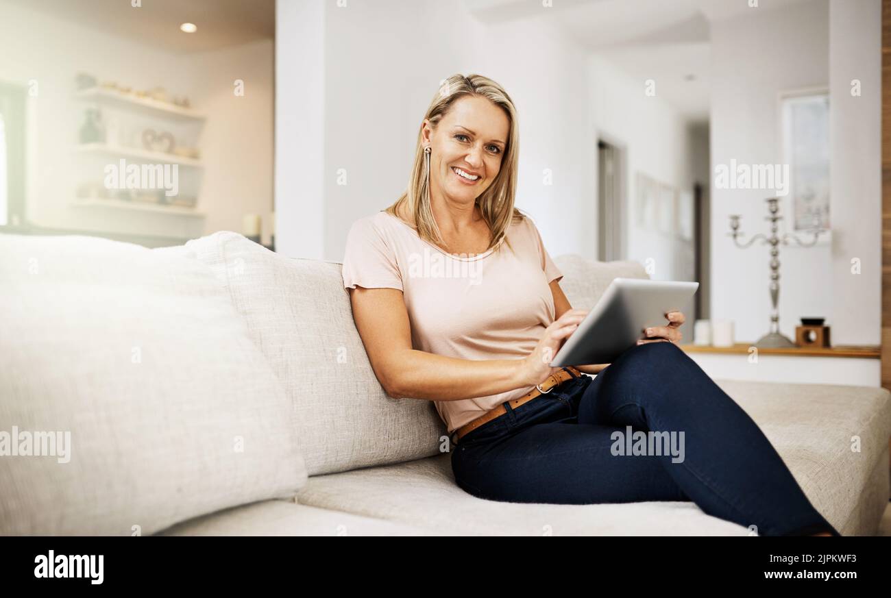 Sorglose und entspannte Frau, die auf einem Tablet stöbert, ihr Wochenende genießt und zu Hause auf dem Sofa sitzt. Porträt einer reifen komfortablen, glücklichen Frau Stockfoto