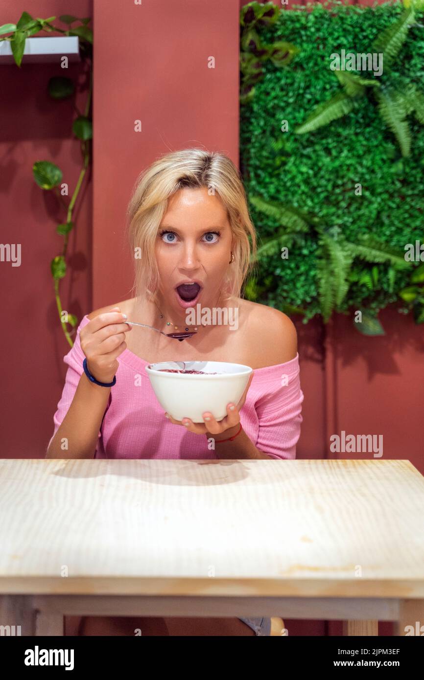 Die Frau schaut schockiert in die Kamera, während sie in einem Café eine acai Smoothie Schale isst. Gesunde Ernährung Konzept. Stockfoto
