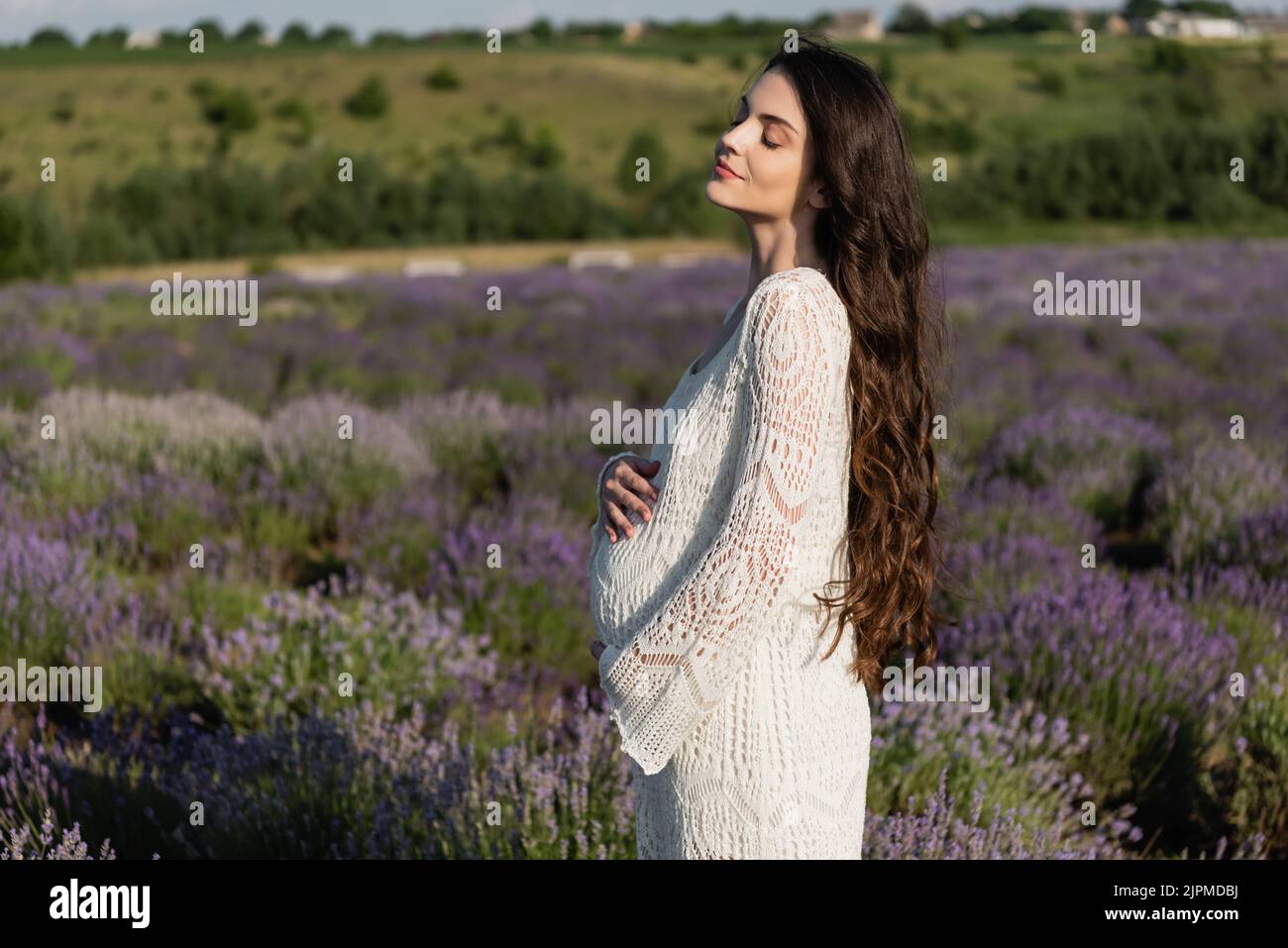 Seitenansicht einer Schwangeren mit langen Haaren, die mit geschlossenen Augen auf einer Wiese mit Lavendel steht Stockfoto