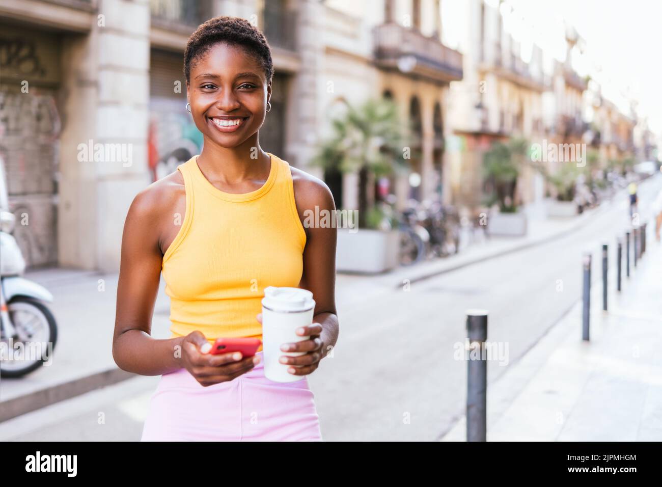 Horizontales Porträt einer positiven afroamerikanischen Frau, die auf der Straße die Kamera anschaut. Sie lächelt und hält einen wiederverwendbaren Kaffeebecher und ein rotes Telefon Stockfoto