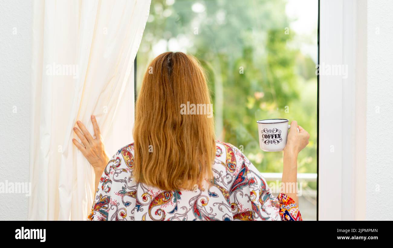 Eine junge Frau schaut mit einer Tasse Kaffee durch das Fenster Stockfoto