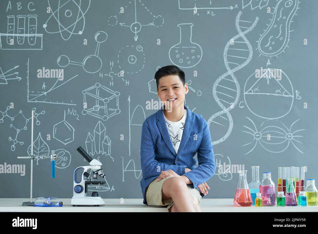 Fröhlicher asiatischer Schüler in Casualwear, der auf dem Schreibtisch sitzt und mit toothy Lächeln für die Fotografie posiert, Inneneinrichtung des Klassenzimmers der modernen Chemie auf dem Hintergrund Stockfoto