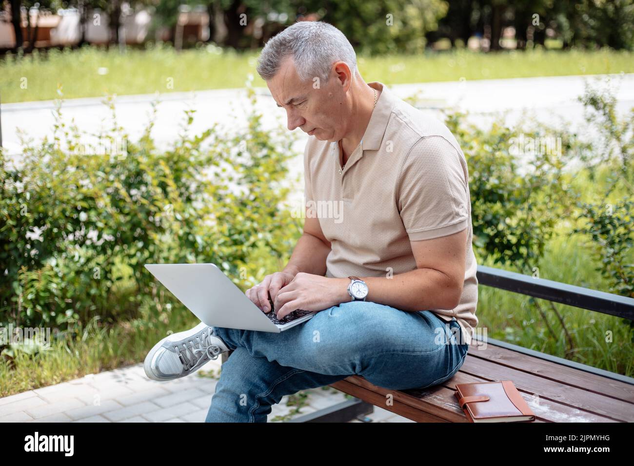 Porträt eines konzentrierten älteren Mannes, der im Sommer auf einer Bank mit gekreuzten Beinen im Park sitzt und am Laptop tippt. Stockfoto