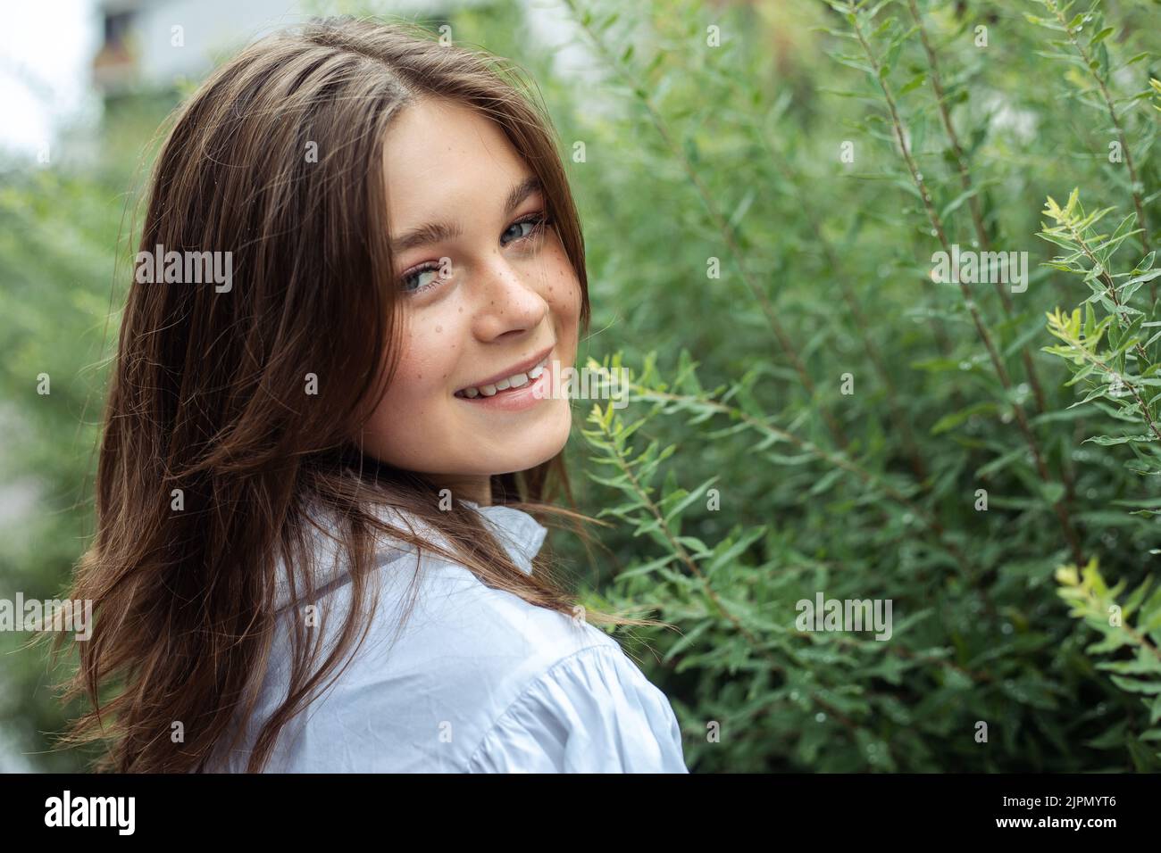Nahaufnahme Portrait hübsches lächelndes Teenager-Mädchen mit grauen Augen und Sommersprossen blicken zurück auf die Kamera in der Nähe von grünem Baum. Speicherplatz kopieren Stockfoto