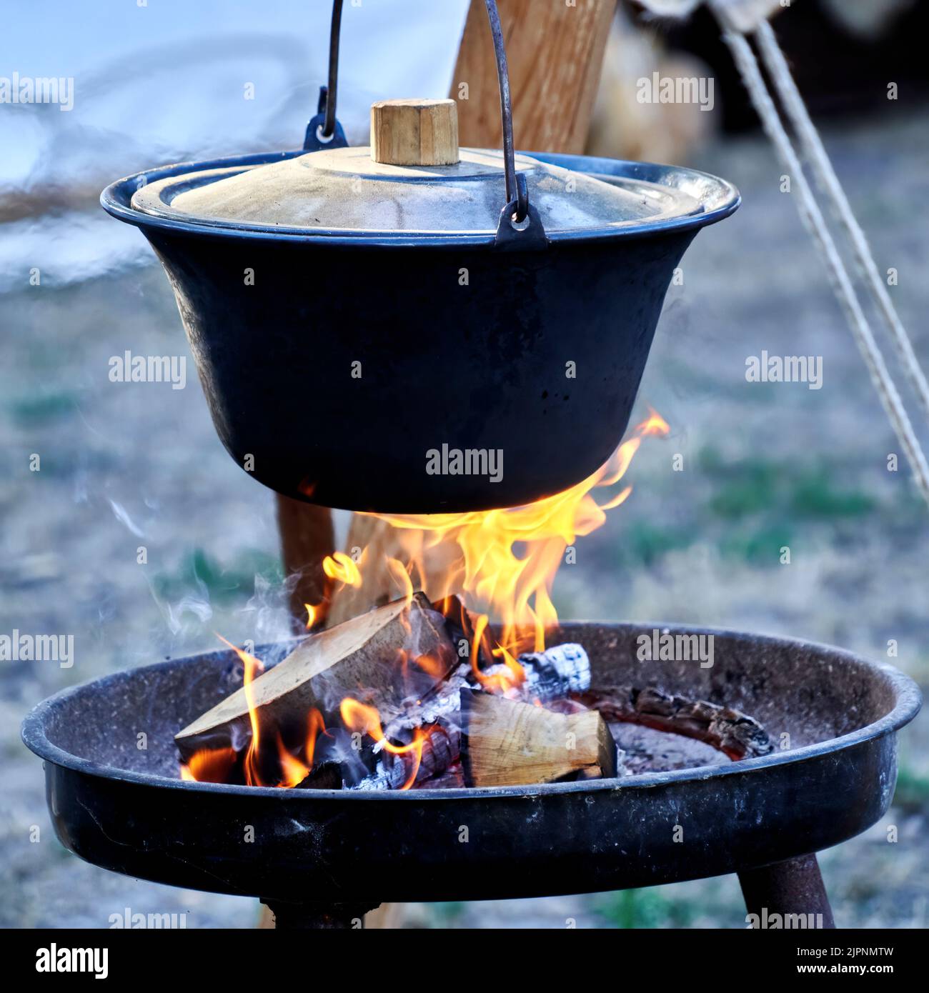 Gusseiserner Topf mit primitivem Deckel hängt über dem offenen Kamin mit brennenden Holzstämmen Stockfoto