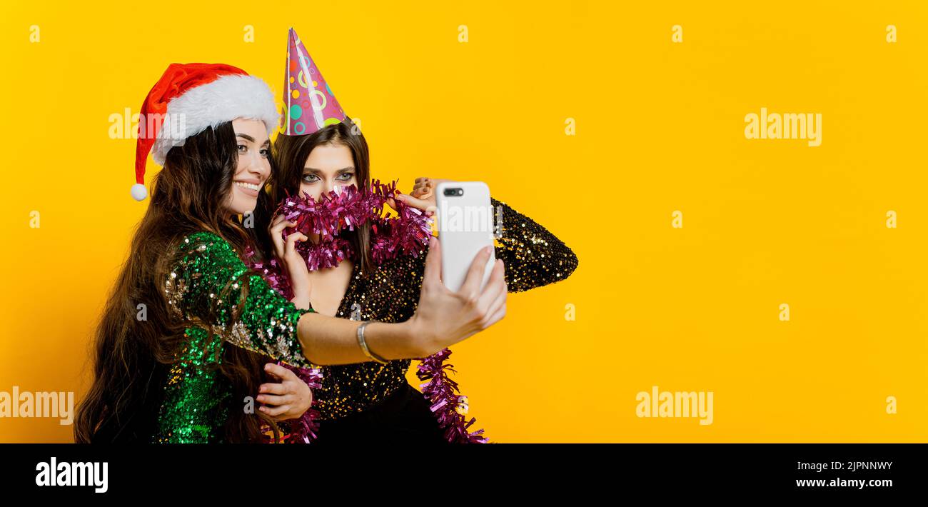 Junge Erwachsene Mädchen 20-25s in einem Partyhut und weihnachtsmannmütze und Lametta machen ein Video oder machen ein Selfie für soziale Netzwerke auf einem gelb isolierten Backgrou Stockfoto