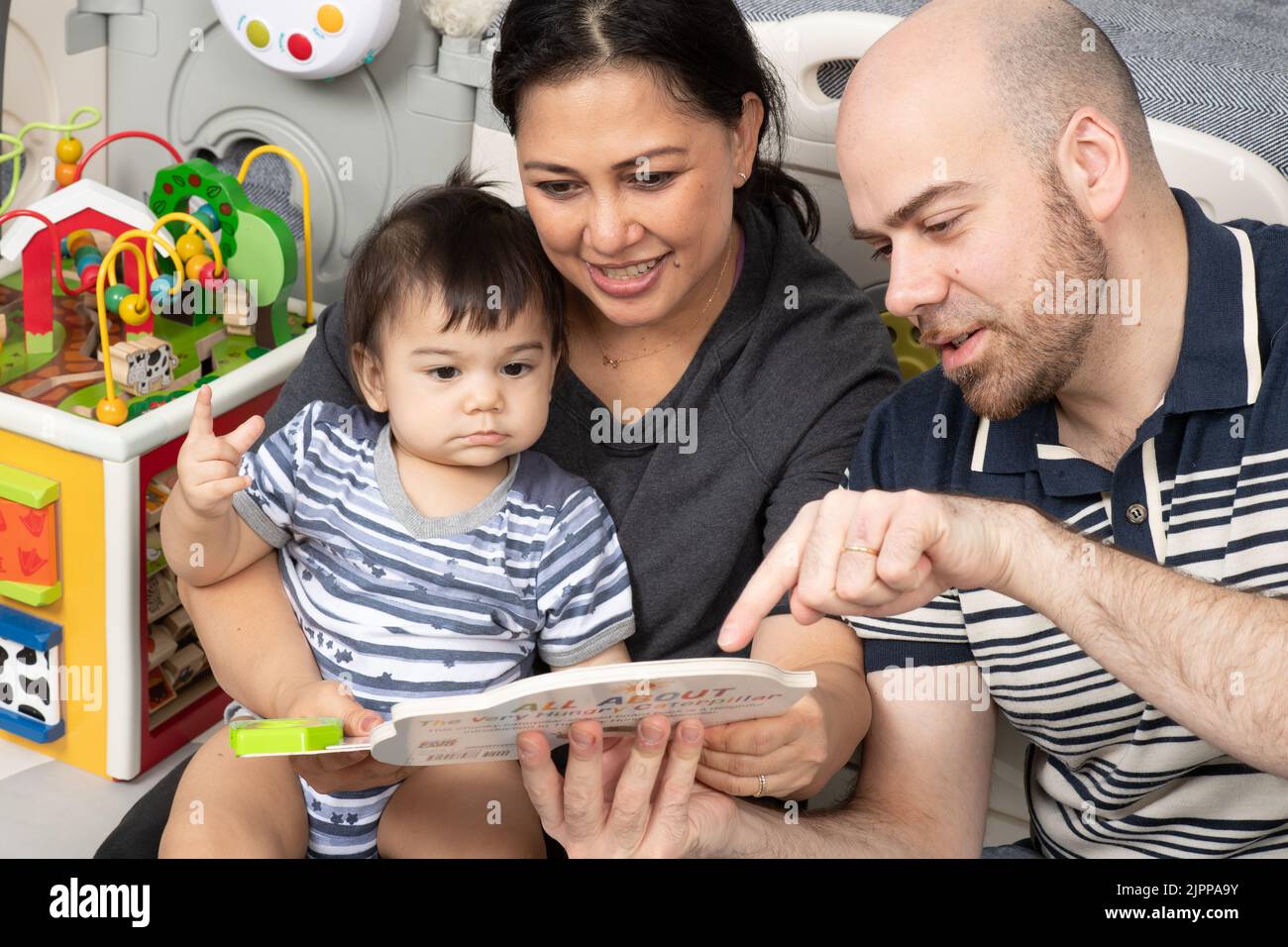 11 Monate alter Junge zu Hause von den Eltern vorgelesen, Vater mit Zeigefinger, um die Aufmerksamkeit auf etwas im Buch zu rufen, Baby hält Zeiger Figur Stockfoto
