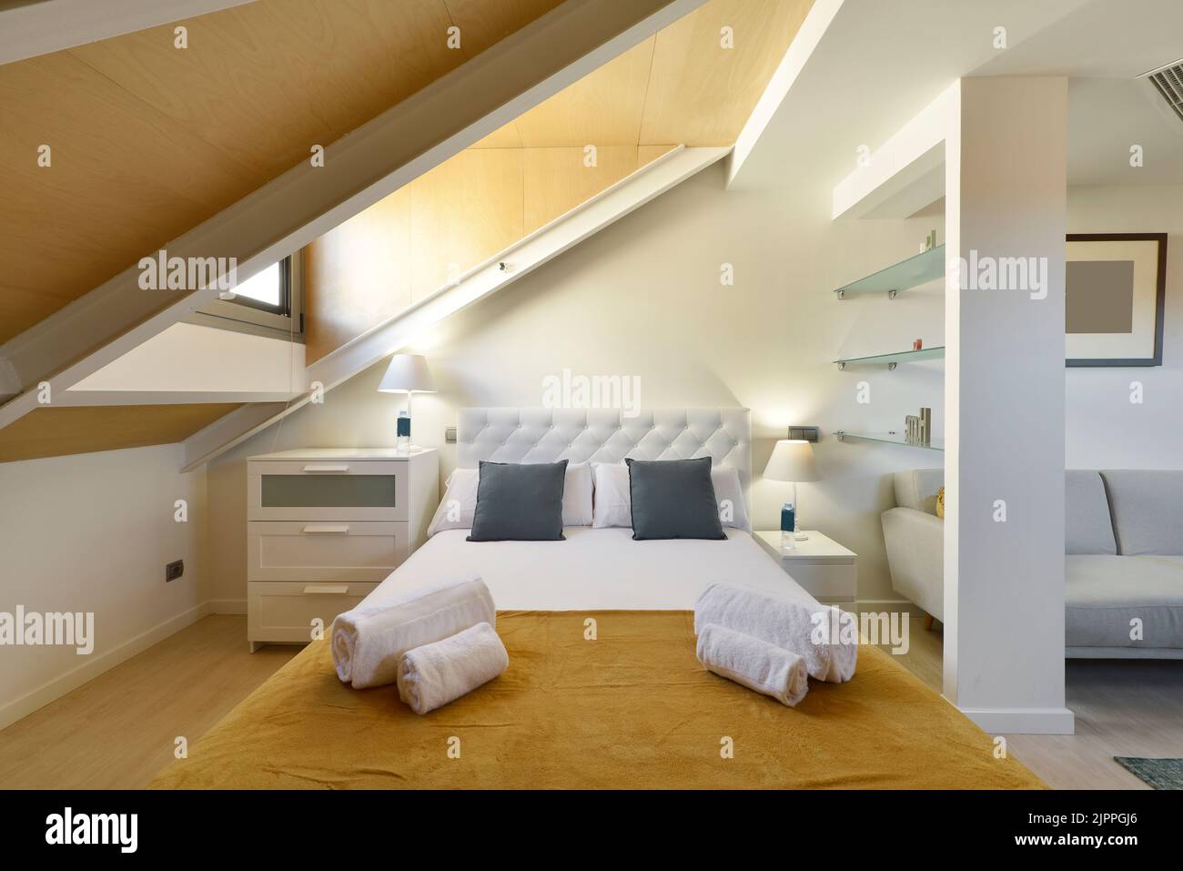 Dekoriertes Schlafzimmer in einem Dachgeschoss-Apartment mit schrägen Decken, Kopfteil aus weißem Stoff, weißen Holzmöbeln und sauberen Handtüchern Stockfoto