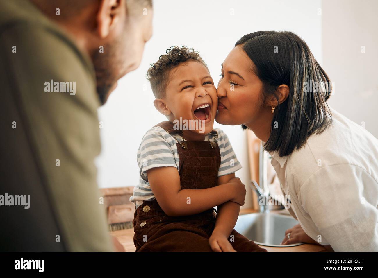Glücklicher Junge, der einen Kuss von der fürsorglichen Mutter bekommt, sich während der Familienzeit zu Hause festmacht und lacht. Junge Eltern teilen sich einen süßen Moment der Elternschaft mit Stockfoto