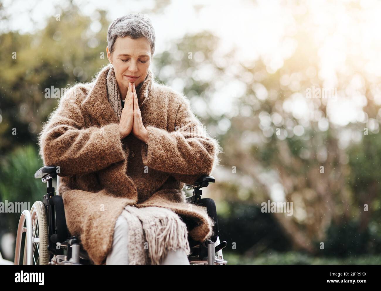 Alles, was ich brauche, ist ein Wunder. Eine ältere Frau hält ihre Hände im Gebet zusammen, während sie draußen in ihrem Rollstuhl sitzt. Stockfoto