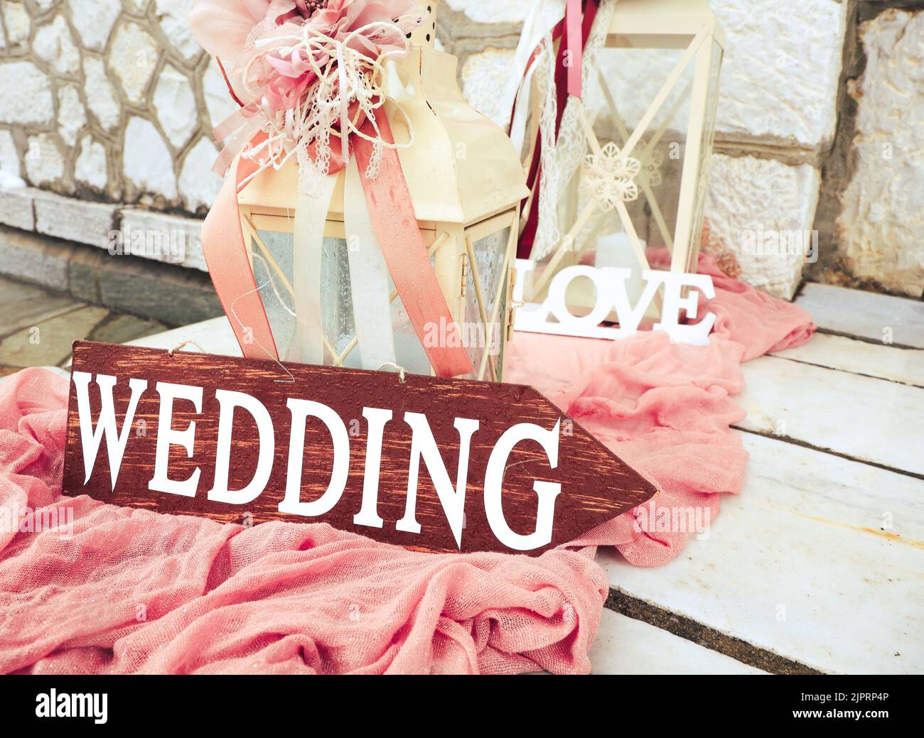 Schöne Hochzeitsdekoration auf steinigen Treppen mit Holzpfeilhochzeit. Große Kerzenlaternen mit rosa Blüten und Bändern auf staubigen rosa Stoff outsid Stockfoto