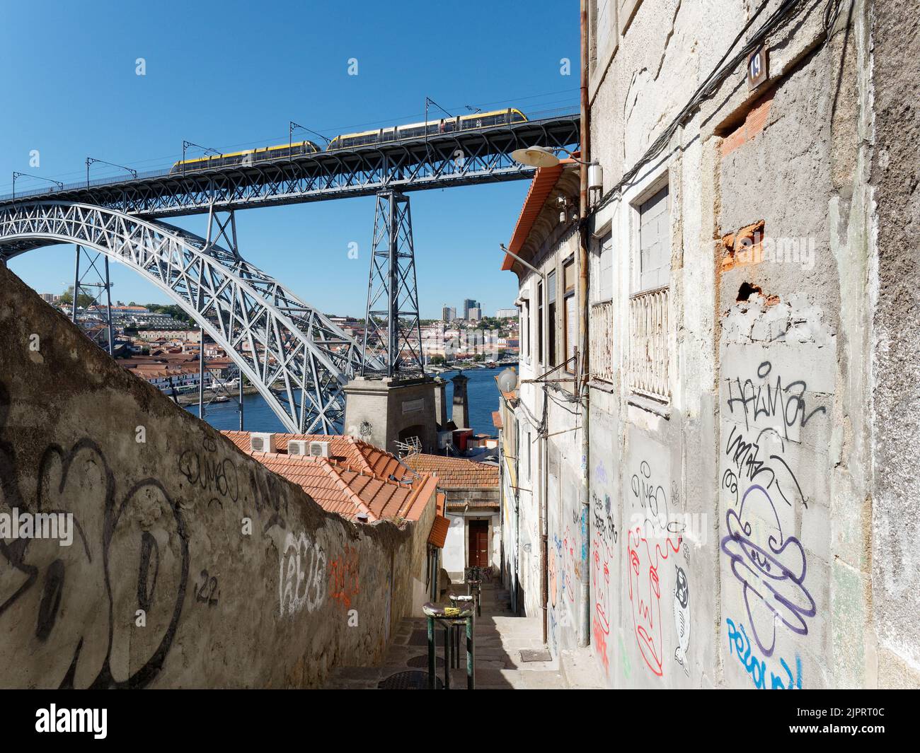 Ein Fußweg führt zum Fluss Douro. Die U-Bahn auf der Luis-I-Brücke ragt hoch und die Mauern sind mit Graffiti bedeckt. Porto, Portugal. Stockfoto