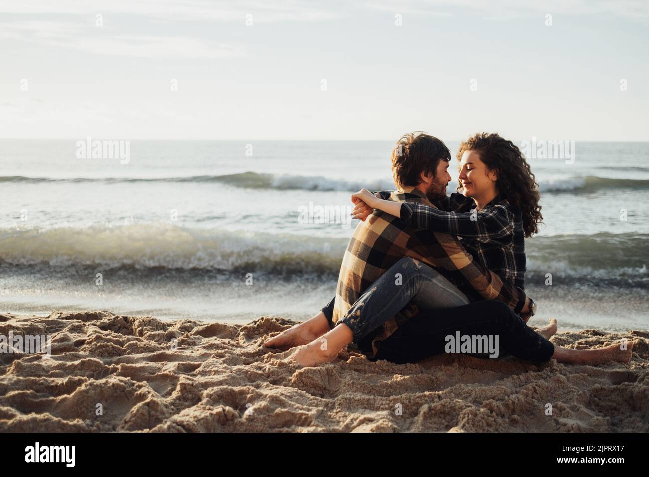 Junges Paar, lockige Frau mit brünetem Mann verlieben sich beim Sonnenaufgang am Meer sitzend und umarmt Stockfoto