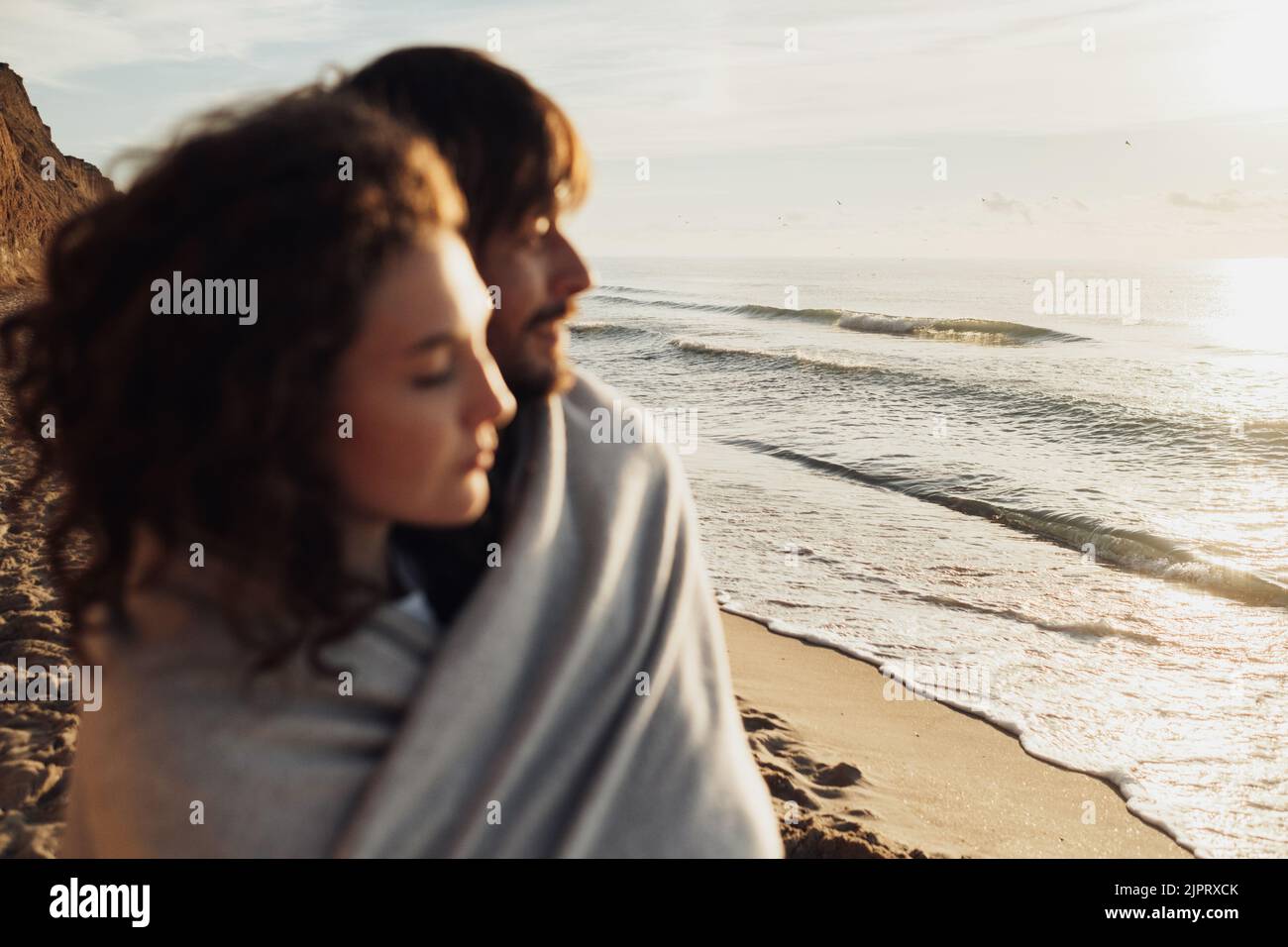 Verschwommenes junges, erwachsenes Paar, eingewickelt in ein Karabinet und am Meer stehend, konzentriert sich auf die Meereswellen Stockfoto