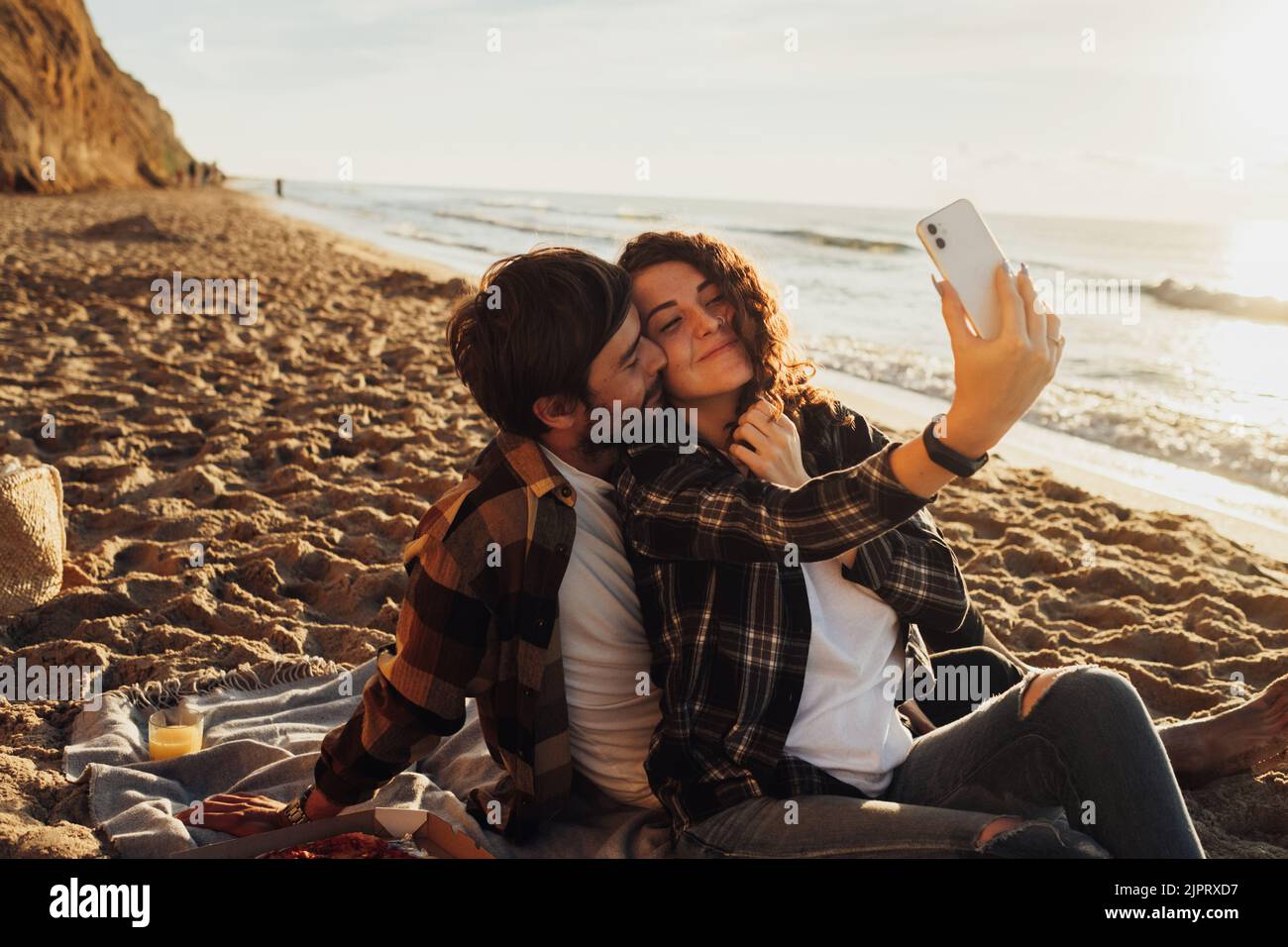 Glückliches junges erwachsenes Paar, das sich verliebt, eine lockige Frau, die zusammen mit ihrem Freund Selfie macht, während sie im Morgengrauen am Meer sitzt Stockfoto