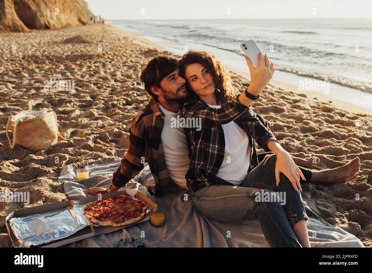 Glückliches junges erwachsenes Paar, Frau und Mann, die gemeinsam Selfie machen, während sie bei Sonnenaufgang ein Picknick am Strand genießen Stockfoto
