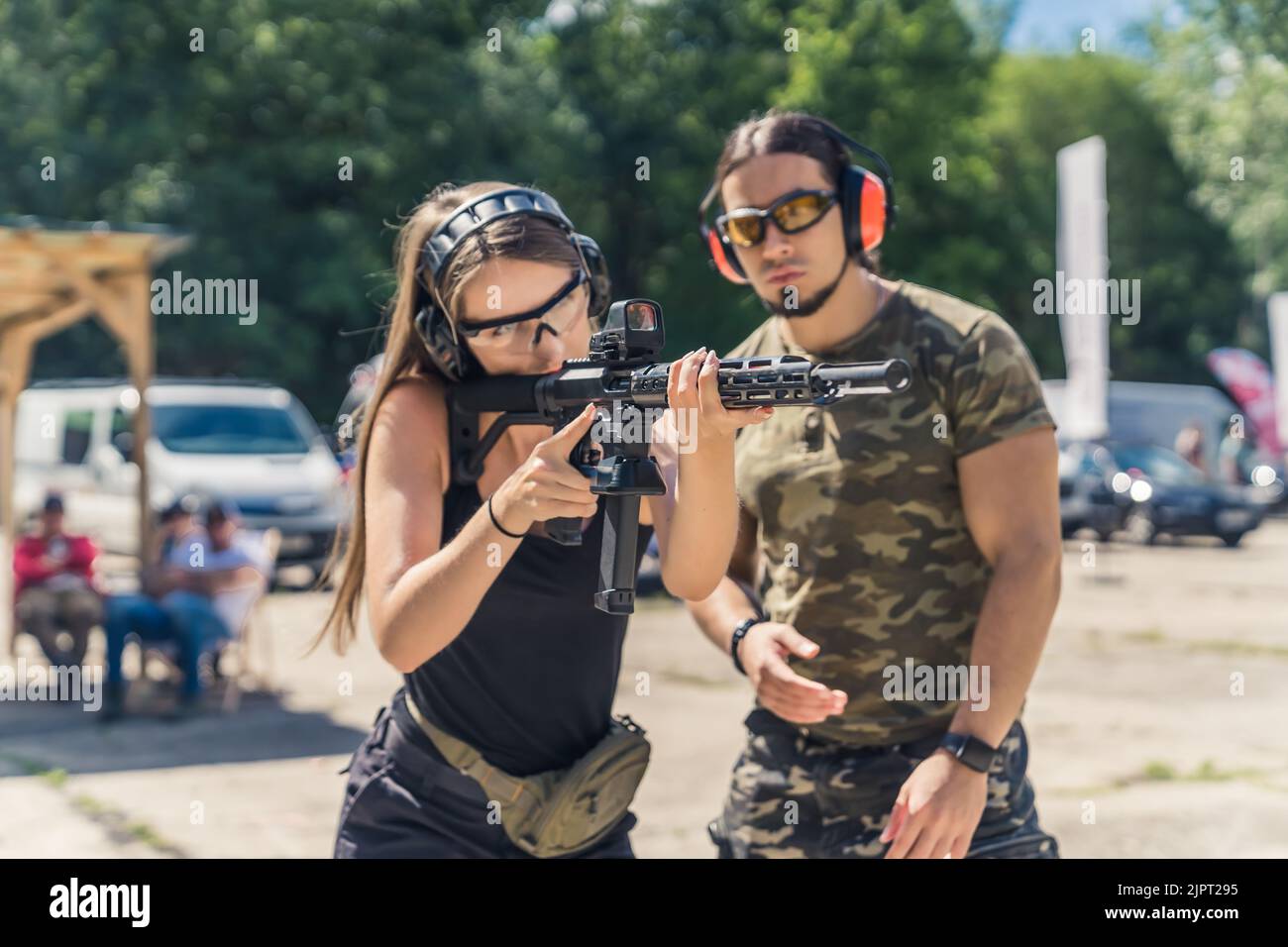 Bärtiger Mann im Camo-T-Shirt, der Frau beim Anpeilen der Maschinenpistole zusieht. Kopfhörer und Schutzbrille. Schusswaffentraining auf dem Schießstand. Horizontale Aufnahme im Freien. Hochwertige Fotos Stockfoto