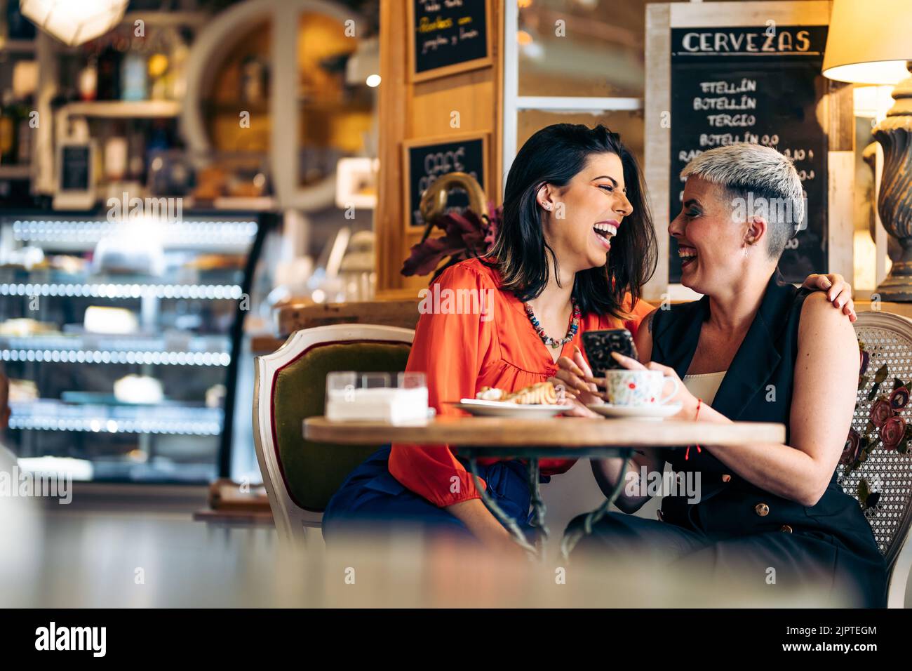 Zwei Erwachsene Freundinnen, die lachen, während sie das Handy betrachten und Kaffee trinken Stockfoto